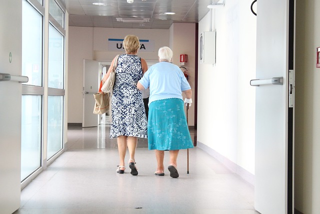 A woman helps a senior woman walk down a hall.